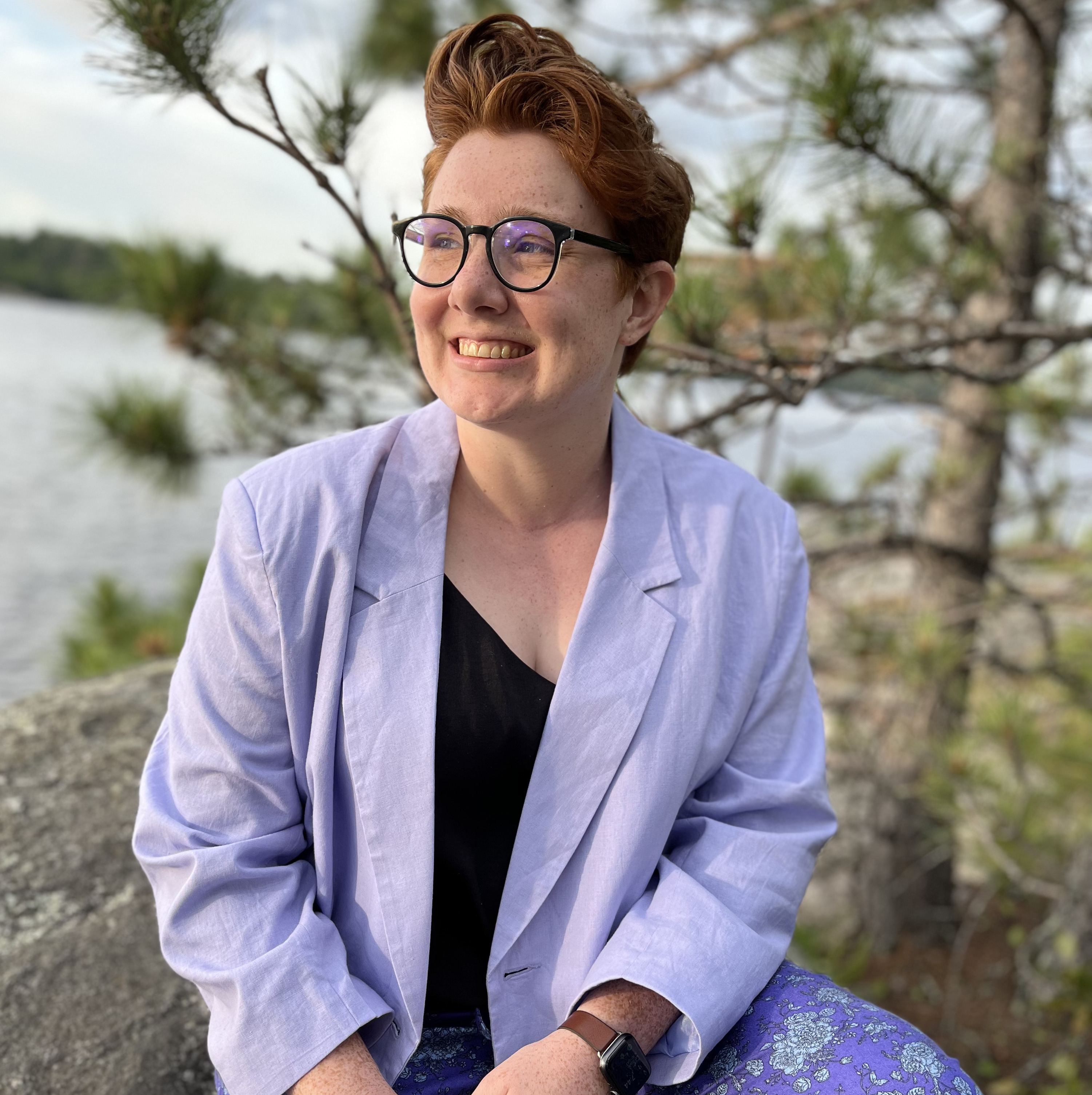 Emma McDonnell, a white woman with short red hair and circular black glassess sits on a rock with a lake blurred in the background behind her, wearing a lavender blazer, black tank top, and floral patterned purple pants. She is not looking directly at the camera, but is smiling looking out.